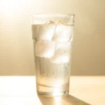 A tall glass of iced water beaded with condensation on a table under bright hazy summer light