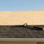 Stacked bundles of asphalt shingles and a tape measure on a partly shingled house roof under a clear sky, no people present