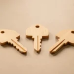 Three plain house keys of different colors resting together on a wooden table next to a small notepad, no text, no numbers, no real person's hands or face