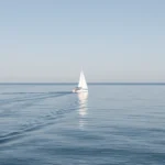 A small sailboat on calm open water with a gentle wake under a clear sky, no text