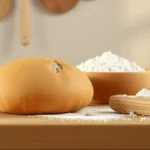A rustic dough loaf beside a wooden bowl of flour and a rolling pin on a floury wooden counter